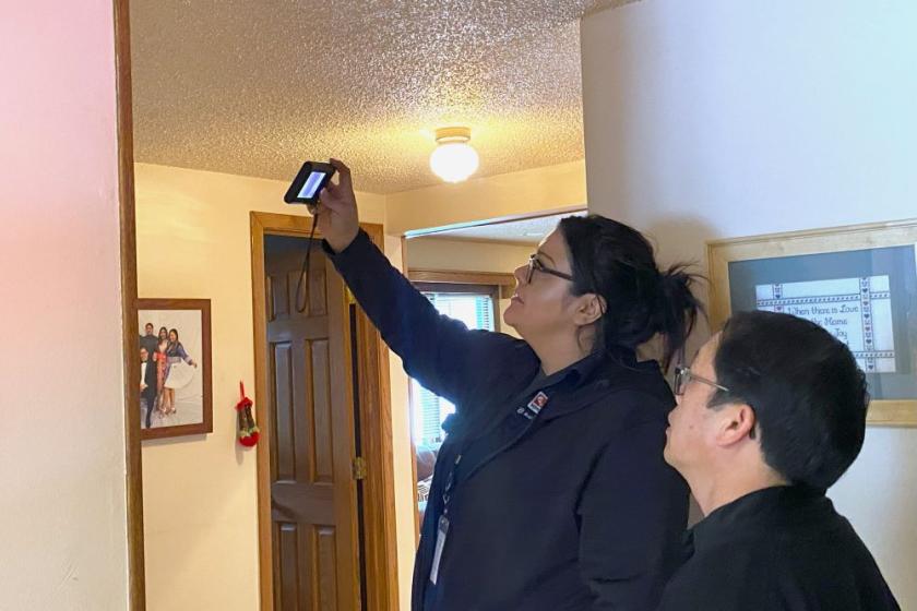 A dark-haired Lakota woman stands in the center of the image, conducting a home energy inspection. She holds up a small electronic device with a screen, pointing it toward the top of a wall. We see her left side. In the right foreground stands a shorter man with black hair and glasses; he is the homeowner. Behind them both is the inside of a home, showing a hallway with a door and a family photo hanging on the wall, slightly out of frame.