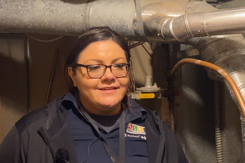 A Lakota woman with her dark hair pulled back, wearing glasses. She is shown in a somewhat close-up shot, from her armpits up. She wears a blue polo shirt with "Home Energy Squad" and "Xcel Energy" logos on the chest. She stands in a basement furnace room with metal furnace equipment above and behind her, and a brown unfinished wall behind her.