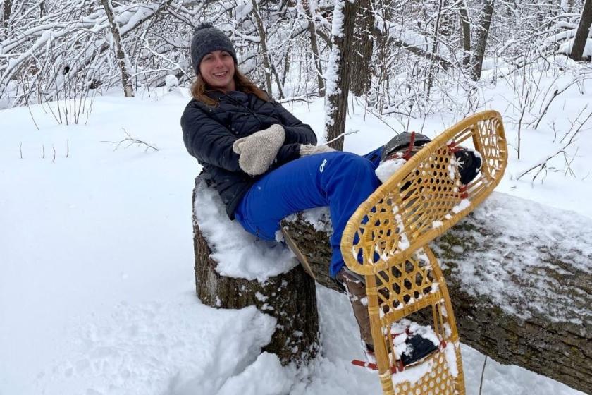  A woman sits peacefully on a broken log in a snow-covered forest clearing, her snow shoes propped up. She is a white woman with dark blonde hair. 