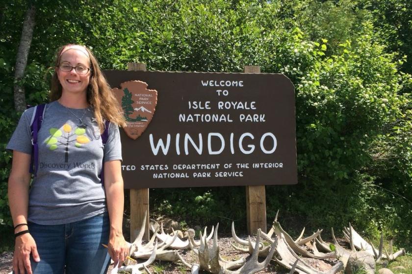 Heidi Auel, a white woman blonde hair, stands outdoors in a wooded area. Behind her is a sign for the Isle Royale National Park Windigo. 