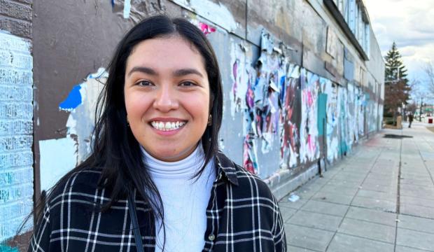 A Latina woman in her early 30's smiles at the camera. She has long black hair and is wearing a plaid blazer and white turtle neck. Behind her is a city sidewalk the exterior of an urban building. It is papered with posters that are pealing off.