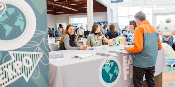 A young woman smiles while sitting behind a check-in desk for the EarthFest Expo. Other people are standing around the desk getting information or talking. 