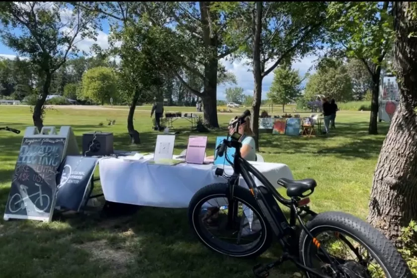 Trees and blue sky in the background.  In the foreground a woman sits at a table with signage sitting on it. An electric bike is in front of the table. 