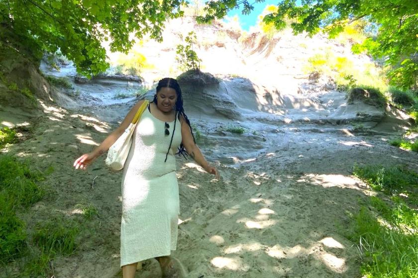 Multiracial woman walking through the sand on a shaded beach, wearing a long white dress with hair in locs. She is surrounded by trees and blue sky is peeking through the trees.