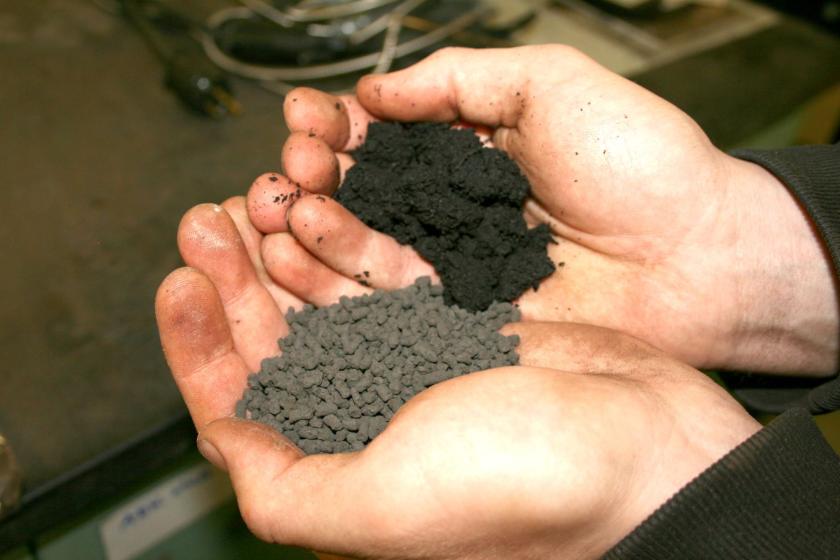 White hands holding two piles of darky earthy material. One hand holds gray pelletes and the second holds a pile of black earth.
