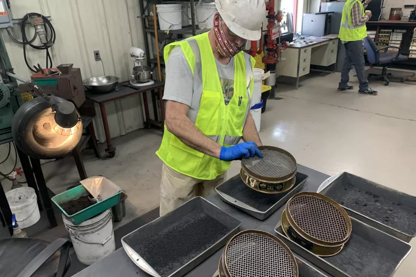 White man wearing a neon yellow vest and white hard hat handling equipment in a manufacturing-style facility. 