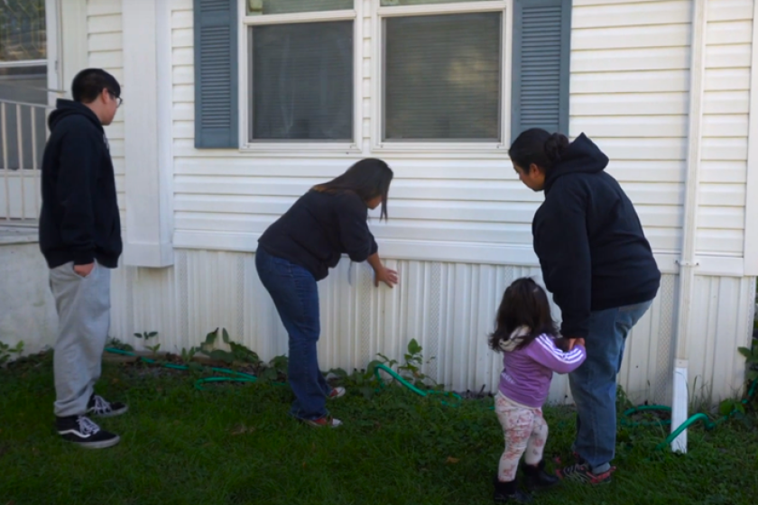 Three adults and one child standing outside a white manufactured home. One woman is pointing at the siding of the home while the other people look on. 