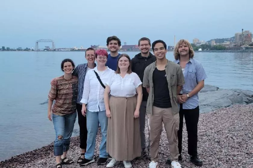 A group of eight people stand on a beach of pebbles with a body of water behind them. In the distance is a Duluth lift bridge. 
