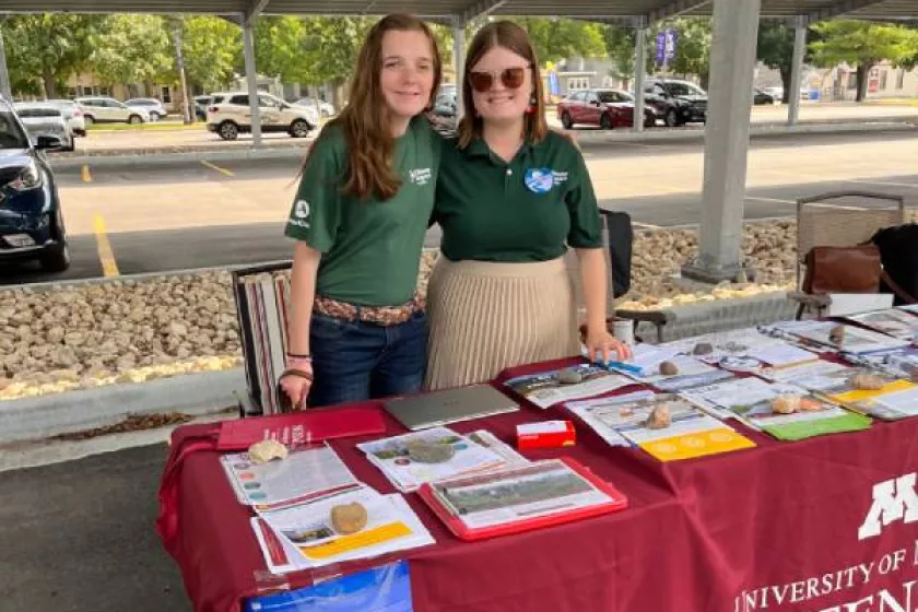 Two young women stand outdoors behind a table. The women stand shoulder-to-shoulder, smiling. 