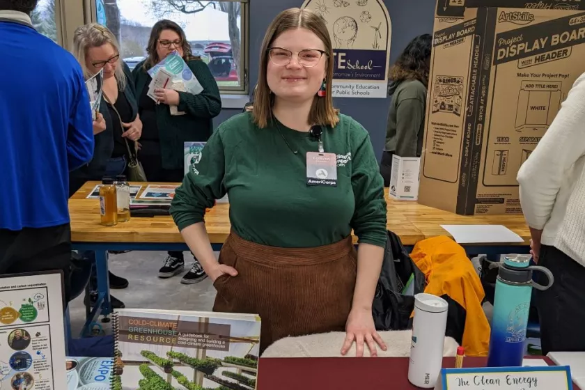 Young white woman stands at a red tabled covered with papers and pamphlets. She wear a green polo and smiles. 