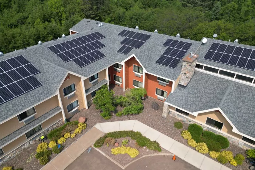 Exterior, overhead view of a modern hotel building.  On the roof, there are rows of solar pa Surrounding the building, there is a sidewalk and open parking spaces. Behind it is a lush green forest. 