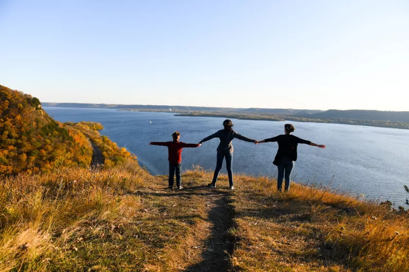 A yellow grassy knoll overlooking a bright blue body of water and a clear blue sky above. On the knoll stands three people, an adult and two children, with their arms stretched horizontally outward. Their features can't be made out but they appear to be looking toward the water. 