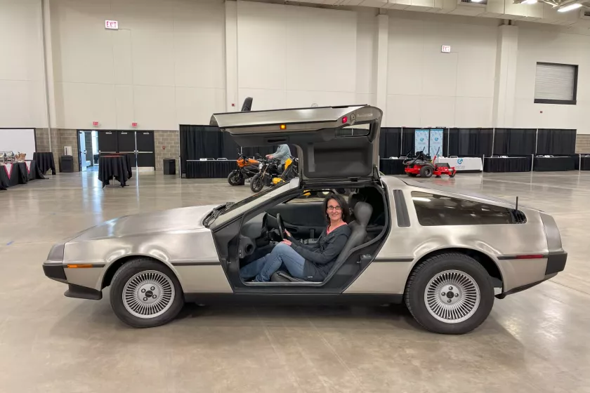 A gray  Delorean car parked in an indoor event space. The drivers door is open toward the ceiling and inside sits a smiling woman