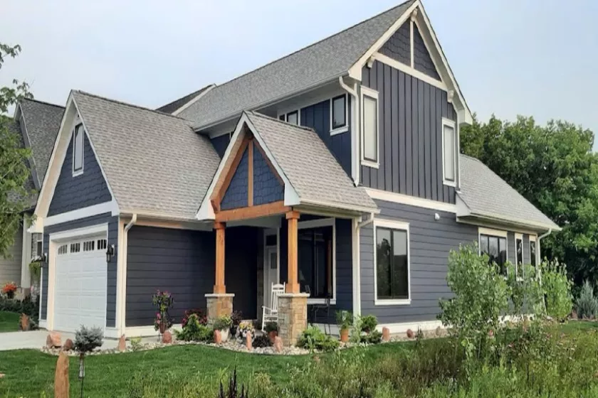 A large blue home with a gray roof and white trim. There is green grass in the foreground and the background shows green trees and blue skies. 