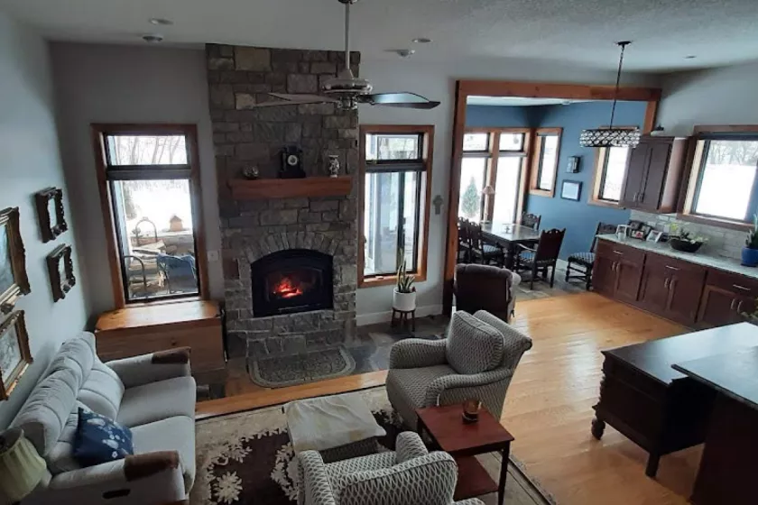 A fish-eye lens shot of a ground floor livingroom. The room has woof floors, a large fireplace, windows drawing in natural light, and cozy furnishing
