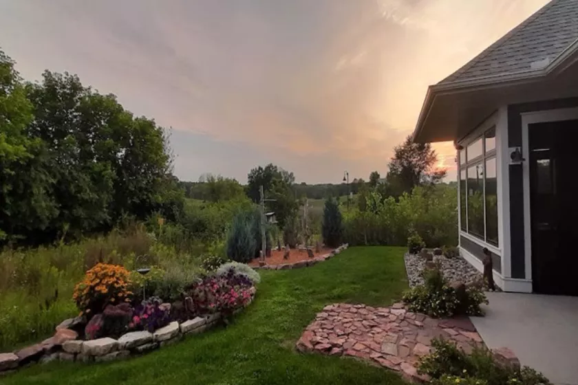 A view from a yard at dusk. An exterior of a home, a rock path,  green grass and trees, and lush garden can be seen. 