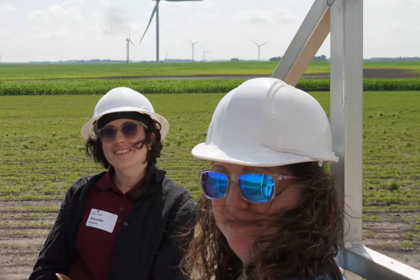 Anna Peterson (right) beside an RSDP colleague as they tour a wind farm. Both women are wearing hardhats, sunglasses, and smiling. Behind them a green field stretches far and wide, and wind turbines can be seen.