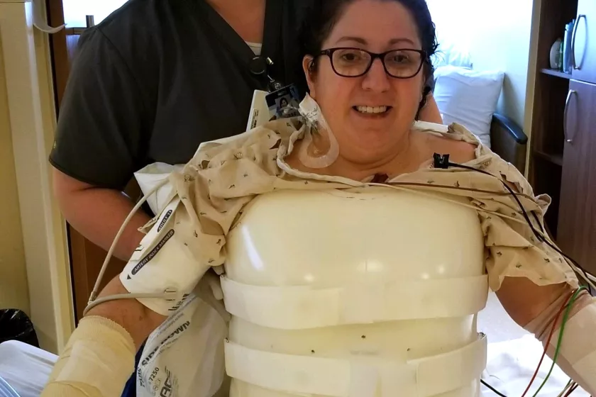  Anna recovering in the hospital. She smiles, wearing a white “turtle shell” body brace. Behind her a medical professional smiles.