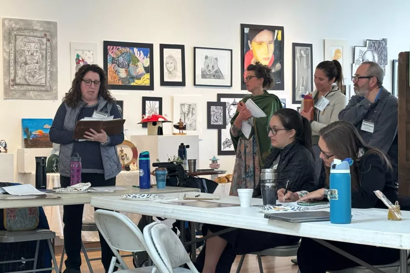 Anna Peterson presents to a group of colleagues in New York Mills. The group is meeting in an art gallery. Anna reads from a binder and her colleagues look on, taking notes and listening intently.