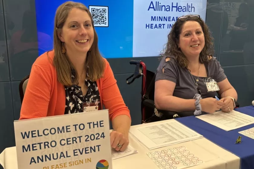 Anna Peterson (right) sitting in a wheelchair behind a table; alongside fellow CERTs regional coordinator, Heidi Auel. A sign at the table welcomes visitors to the “Metro CERT 2024 Annual Event.” Both women are smiling, welcoming guests to the event. 