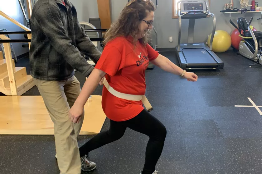 Peterson taking part in physical therapy. She stands in a lunge position while Trevor Roppel, DPT of Choice Therapy stands behind her, helping support her. Exercise equipment stands in the background. 