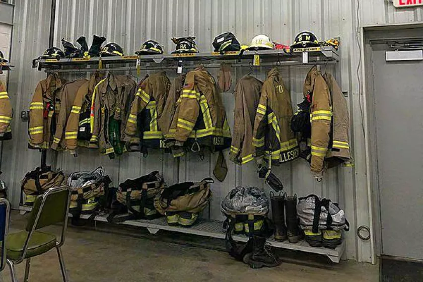The inside of Comfrey’s fire hall. A row of fire department jackets and gear are stored on shelves mounted on a metal wall. High above, a LED lighting is mounted on the ceiling.