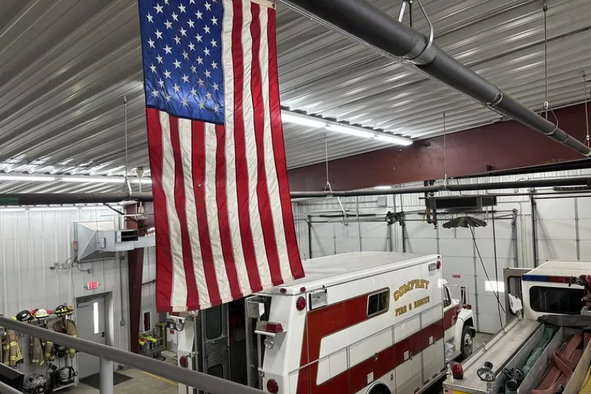 The inside of Comfrey’s fire hall. An American flag hangs from the ceiling in the foreground. Behind it, rows of lights are visible on the ceiling. Below, a Comfrey Fire & Rescue Truck and a fire truck are parked.