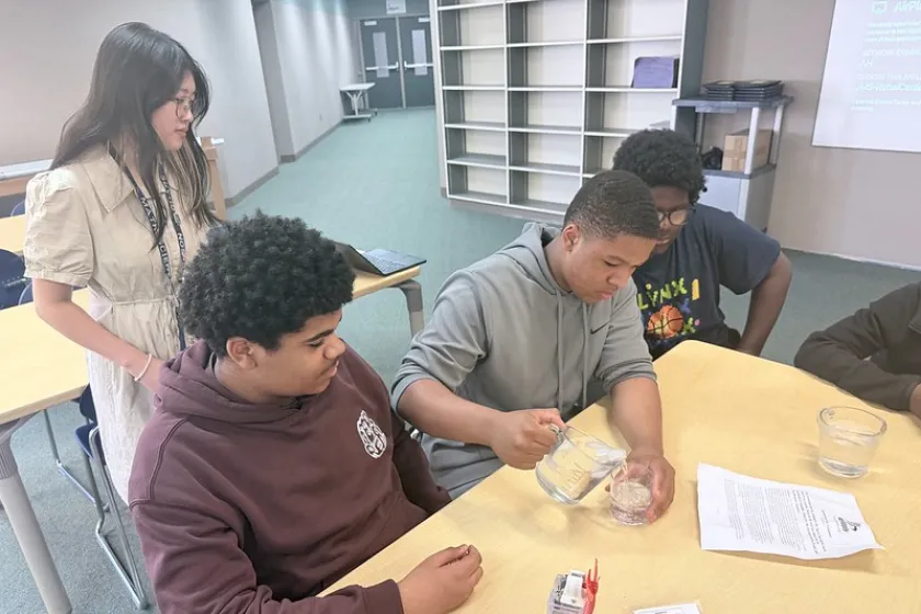 Three students measure water into a cup in a classroom as a teacher watches over.