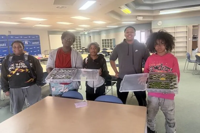  Five youth hold up two boxes of small plants growing from disposable condiment paper cups in a classroom.