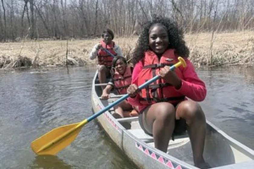 Three young Black women smiling in a canoe on the water. 2026.