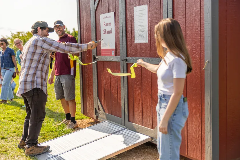 Two people hold a ribbon in front of barn doors while a third man cuts it.