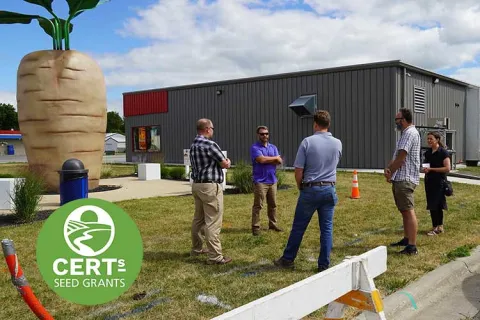 Group of men standing on a lawn, next to a large grey building in the background, and a giant parsnip statue. 