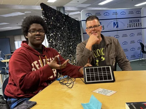 : A middle aged white man sits next to a Black young man showing a phone plugged into a small solar panel. 2026
