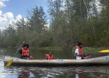  Three young Black men waving paddles on their canoe on the water. 2026.