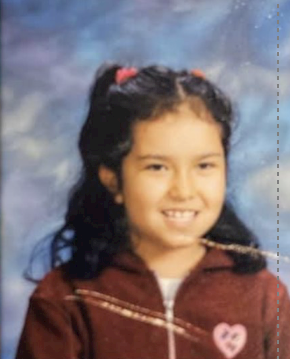 A young Latina girl smiles at the camera. She is wearing a maroon colored zip up track jacket and has her hair styled with two pink pony tails on top. Behind her is a blue backdrop. 