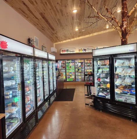 The inside on a convenience store. Three large, standup coolers can be seen. They shine bright white light and are stocked with items like pop, bags of frozen food, and other grocery items. The ceiling, walls and floor and all warm shades of brown and look freshly constructed. A large, decorative birch tree trunk and bare limbs can be seen above the coolers, reaching toward the ceiling. 