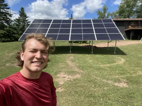 A young man takes an outdoor selfie. In the foreground, the white, blonde man wear a red shirt and squints into the sun with a big smile. Behind him a large solar panel stands in a grassy area. A home with a deck and blue skies can be seen in the background.