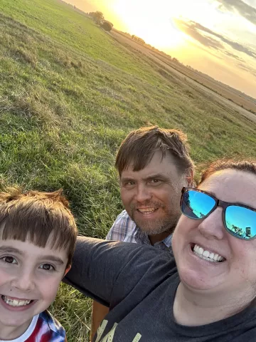 Peterson (right) stands in a grassy farm field with her husband and youngest child. The group smiles. In the background, the sky is an orange hue and the sun hangs low on their last night of harvest. 