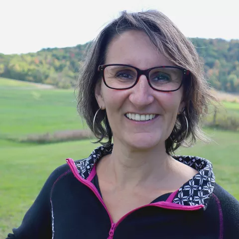 A close up headshot of a woman. She appears white with chin-length brown hair. She has a big smile. She wears hoop earrings and glasses. The background is blurred but a large grassy knoll and trees can be seen stretching out in the distance. 