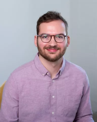Close up photo of man smiling. He appears to be white with brown hair and a beard. He is wearing glasses and a light red shirt. 