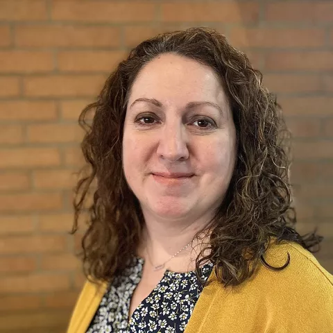 Professional headshot of Anna Peterson, Northwest CERT Regional Coordinator. She is smiling, wearing a yellow sweater and a floral black and white blouse. The background is a brick wall.