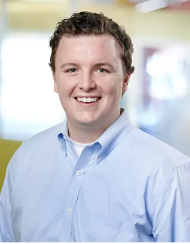  Bryn Shank, a white-presenting man with blue eyes and curly brown hair, smiles in a professional photo.