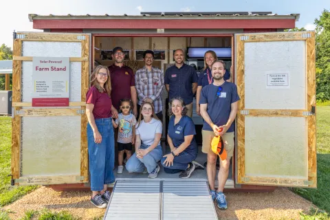 Nine people stand just inside a farm stand shed.