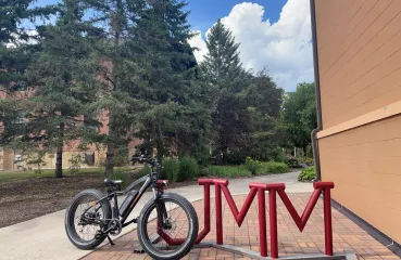 Blue sky and pinetrees in background. In the foreground an electric bike sits against a bike rack.