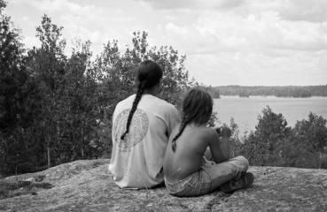 Black and white image. A man with a braid is sitting on a rock structure. He sits close to a boy with a braid. Their backs face us as they overlook a cliff and trees. 