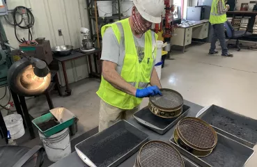 White man wearing a neon yellow vest and white hard hat handling equipment in a manufacturing-style facility. 