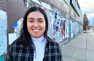 A Latina woman in her early 30's smiles at the camera. She has long black hair and is wearing a plaid blazer and white turtle neck. Behind her is a city sidewalk the exterior of an urban building. It is papered with posters that are pealing off.