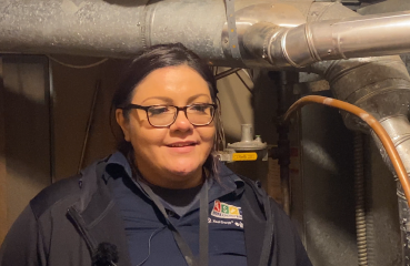 A Lakota woman smiles at the camera, with her dark hair pulled back, wearing glasses. She is shown in a somewhat close-up shot, from her armpits up. She wears a blue polo shirt with "Home Energy Squad" and "Xcel Energy" logos on the chest. She stands in a basement furnace room with metal furnace equipment above and behind her, and a brown unfinished wall behind her.