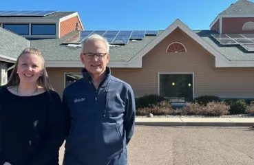 White woman and man stand shoulder to shoulder outside. Blue sky and hotel in background.