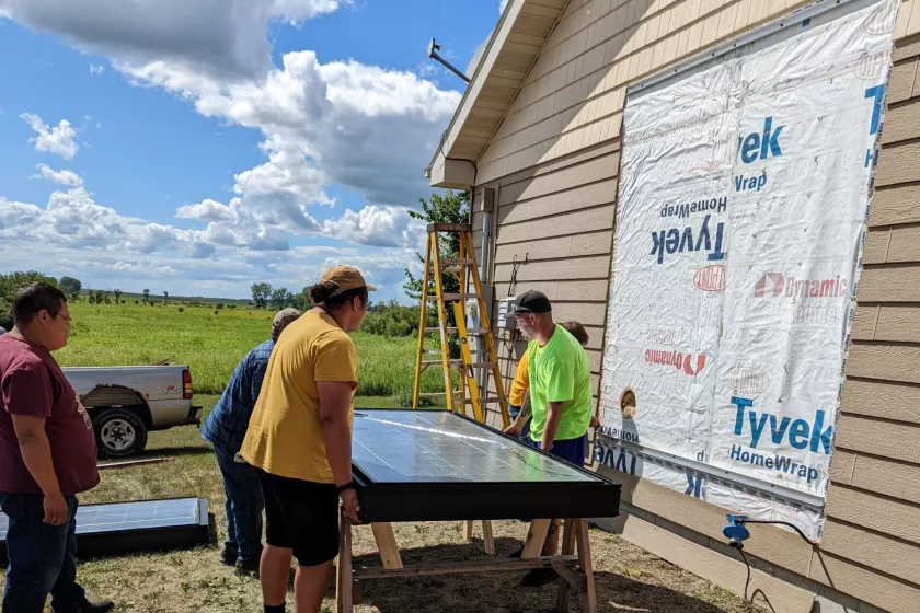 A group of people standing outside near a home. The side of the home has siding cut out, the people are standing near a solar thermal panel. 